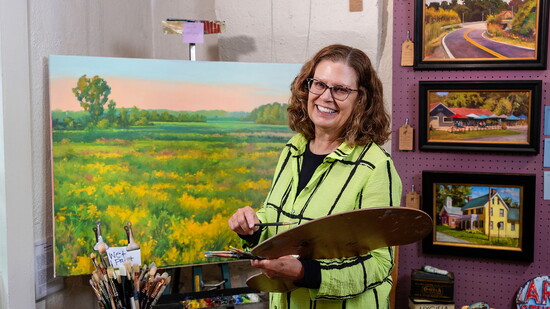Gloria at her easel, photographed from an angle that shows both her focus on the work and the painting in progress.