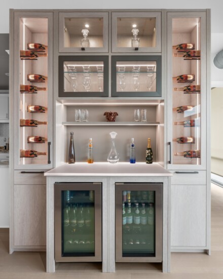 A wine bar features integrated lighting with wine pegs to display favorite bottles. The countertop is Chroma, a back-lit resin; the back panel above the counter