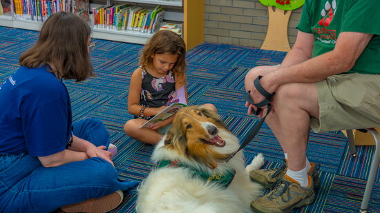 Beginning reader Stella shares her book with Eoin, his handler Ken Wegenhart & Madeira Library Customer Specialist Isabella Randolph.