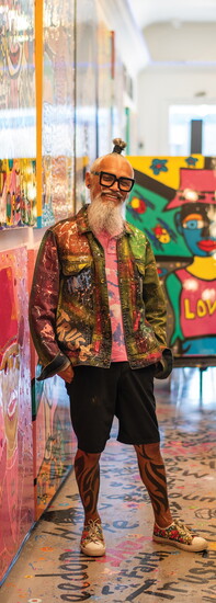TAVi's personal style is as colorful as his art, from his paint-splattered jacket and bold tattoos to his signature white beard and oversized glasses.