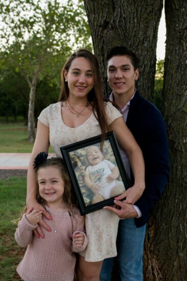 Kristen and Jacoby Gonzales (shown with daughter Emmaline) hold up a photo of their infant daughter, Penelope, who died of SIDS in 2016. (Photo provided)