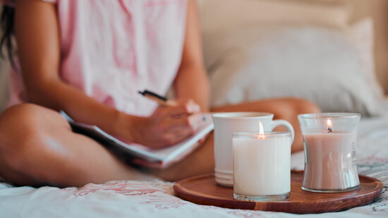 A person journaling in bed with candles and a warm drink.