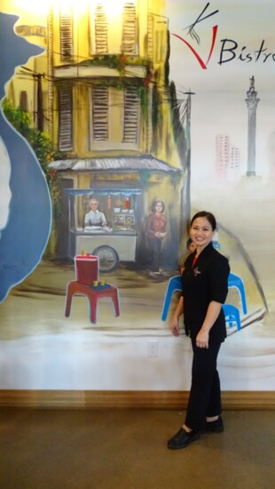 Julie Vo standing next to mural of her family on the wall in V-Bistro. 