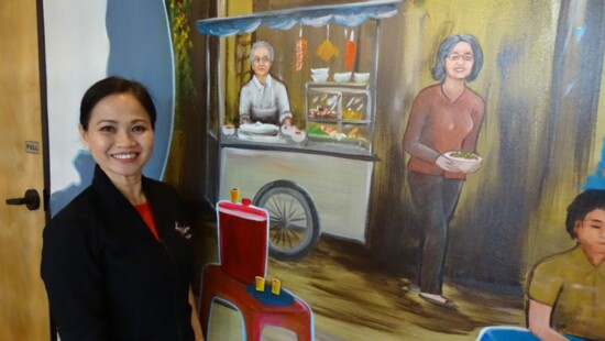 Julie Vo, Owner/GM, stands beside the mural of her family serving from a food cart in Vietnam. 