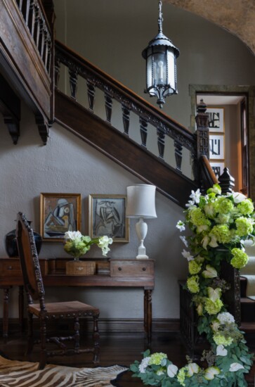 Eclectic artwork and fresh flowers are spotlighted by natural light from leaded glass windows anchoring a carved staircase.