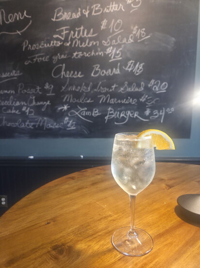 A sparkling cocktail in front of a menu board in the bar area at Près Des Prés. Photo Credit: Jennifer Waldera