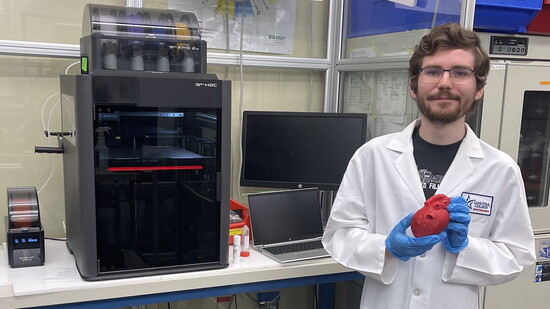 Samuel Hopkins standing near a state-of-the-art 3D printer with a model of a patient's heart printed from a CT scan.
