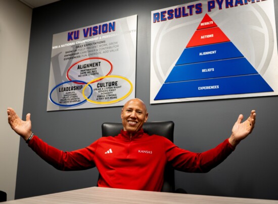 Coach Q in his office showing the posters that highlight the KU baskeball team goals.