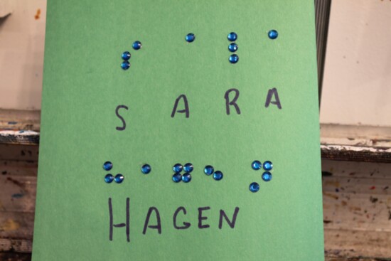 Image Description: the name Sara Hagen written in Braille using blue rhinestones on green construction paper