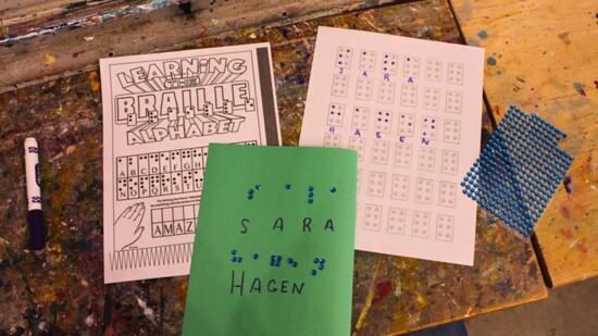 Image Description: art desk with an alphabetical Braille chart, a practice Braille sheet, and the full name of Sara Hagen spelled out in Braille