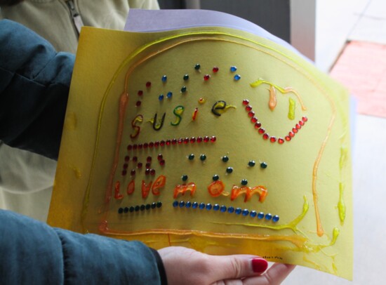Image Description:  Someone's finished rhinestone Braille project, their mom holding out the piece of yellow construction paper with their name on it in Braille